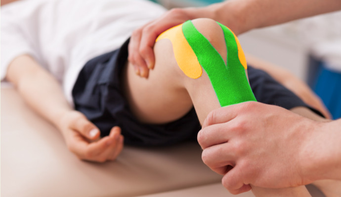 Auf einem Knie ist Kinesio Tape angebracht und zwei Haende eines Physiotherapeuten korrigieren die Haltung des Beins.